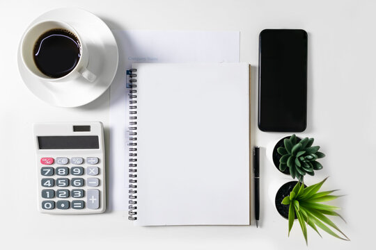 Blank white notebook with empty space in the middle. On the white table are a calculator and a mobile phone and a coffee cup, for mockup. Empty space for text, graphics work.