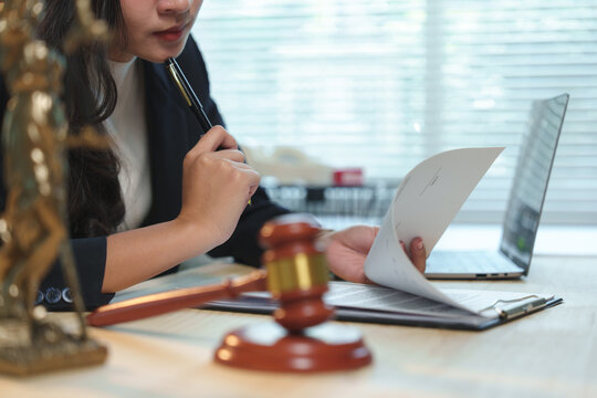 Female lawyer is carefully reviewing legal documents in her office, holding a pen and contemplating the next steps in a case, with a gavel and statue of justice symbolizing law and order