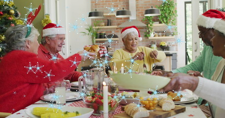 Image of snow falling over senior diverse friends in santa hats at christmas meal table