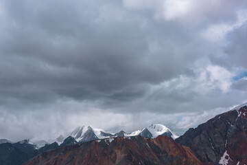 Dark atmospheric alpine landscape with sharp rocky ridge and snowy peaked tops in low heavy lead gray cloudy sky. Rock mountain wall and snow-capped pointy peaks in bleak dramatic bad rainy weather.