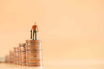 Retired elderly model holding a cane standing on gold coins with growing value on the brown background in the office, Saving money for retirement and planning in the future concept.
