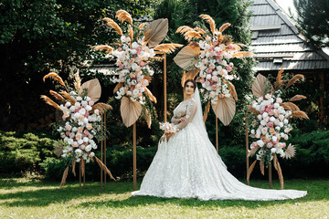 bride in a wedding dress standing against the backdrop of a floral arch. White dress with a long...