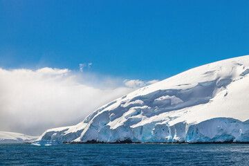 Antarctic peninsula covered in deep snow. Ocean in foreground; blue sky and clouds behind. 
