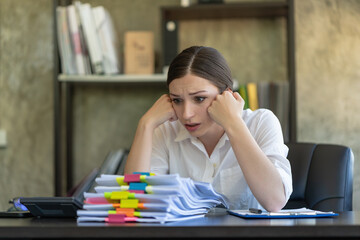 A stressed woman sitting at a desk in an office surrounded by paperwork.
