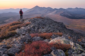 A hiker stands atop the mountain peak, overlooking a beautiful scene at sunrise. The rugged terrain is covered with rocks and shrubs, creating an adventurous atmosphere. In the distance