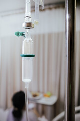 Closeup View of Intravenous Infusion Drip with Female Patient Sitting on Hospital Bed on the Background