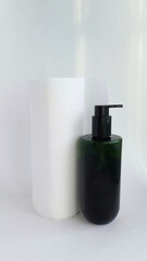 A dark green bottle with a dispenser and a roll of paper towels on a white background. Cosmetic products for skin and hair care. Liquid soap.