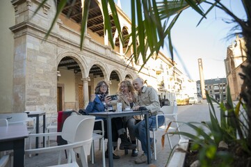 Senior friends taking a cheerful selfie at an outdoor café in a historic sunlit plaza surrounded...