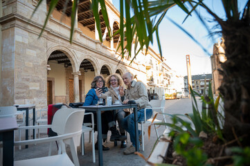 Three senior friends enjoying coffee and taking a selfie at an outdoor café in the main square of a picturesque Spanish village