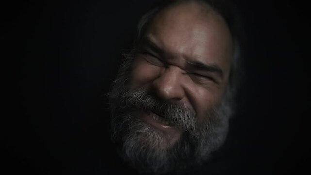 Closeup portrait of a mad, angry screaming bearded old man making faces on a black background. Sticking out tongue at camera. Fury, insanity, anger, madness topics. Slow motion video.