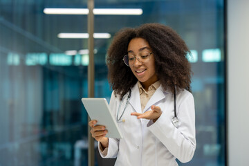 Online doctor consultation, young female doctor in white medical coat uses tablet computer for video call, remotely consults patient, smiling happily inside clinic office.