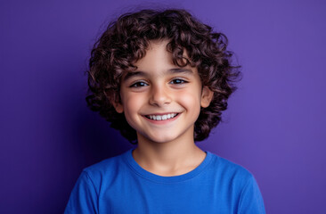 A joyful boy with curly hair is smiling widely, showcasing his happiness. The vibrant purple background adds a playful touch to his cheerful expression, capturing a moment of fun.