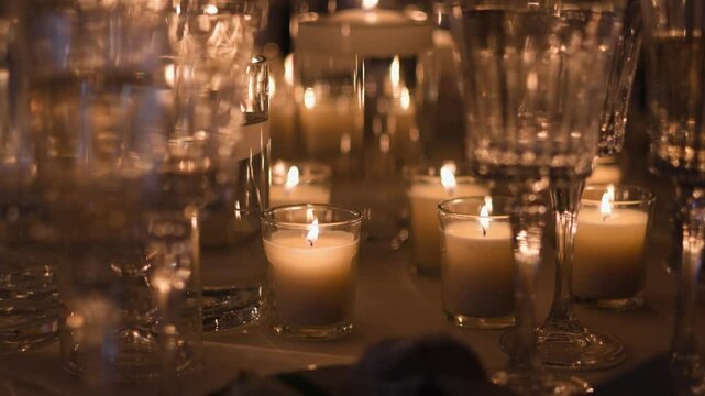 A collection of lit candles in glass holders, surrounded by reflective glassware. The dim lighting creates a cozy and intimate atmosphere, ideal for relaxation, events, or romantic settings.