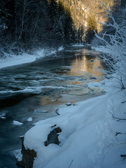 Sunlit River Cutting Through A Frosty Winter Forest: A Tranquil Harmony Of Snow, Ice, And Golden Light