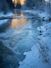 Sunlit River Cutting Through A Frosty Winter Forest: A Tranquil Harmony Of Snow, Ice, And Golden Light