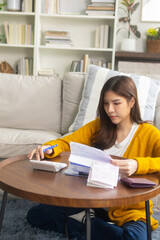 A focused young Asian woman sits at a desk in her living room at home, using a calculator to plan her monthly budget plan, ensure her bills, and focus on managing her expenses and financial goals.