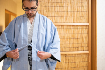 Man wearing traditional Japanese yukata indoors, adjusting the fabric tie. Bamboo screen in...