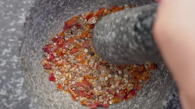 Hand with a pestle is grinding and mixing various kinds of spices, peppers and sea salt in the grey stone mortar. High quality 4k footage