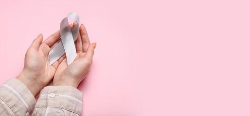Hands of woman holding brain cancer awareness ribbon on pink background with space for text