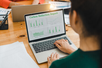person analyzing carbon reduction report on laptop screen, with green graphs and charts displayed, while sitting at wooden desk with documents and colleagues in background