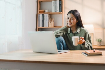 young woman sits at wooden desk with laptop, holding coffee mug and smiling, enjoying relaxed moment in bright home office with bookshelves and natural light