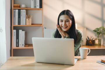 cheerful woman smiling while working on laptop at wooden desk with coffee mug, bookshelves, and natural light in background