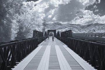 Obraz premium Infrared photography, a long, narrow bridge extending into the distance with wooden planks forming the pathway at Black bridge, Lampang, Thailand