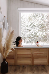 A young woman is relaxing lying in the bathtub in a cozy bathroom with panoramic windows overlooking a snowy forest in a country house in winter. Girl takes water treatments at a spa. Selective focus 