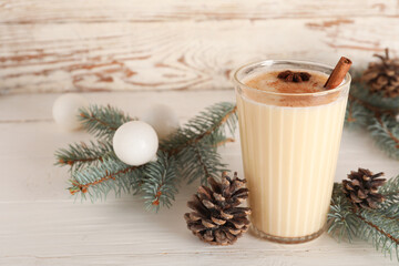 Glass of tasty eggnog cocktail with fir branches, Christmas balls and pine cones on white wooden background