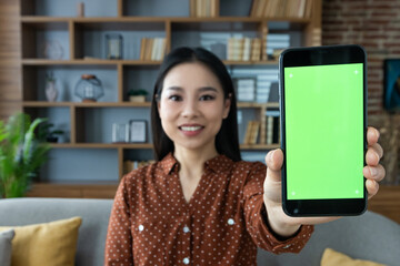 Asian woman in casual attire shows smartphone with vibrant green screen at home. Focus on phone...