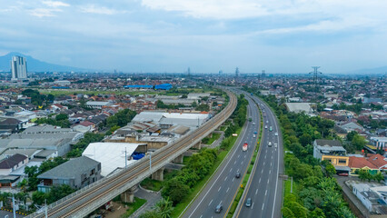 Drone photo aerial view of Bandung Cileunyi highway toll road traffic with Jakarta Bandung High Speed Railway surrounded by  residential areas, industrial areas and warehouses in cloudy afternoon