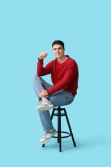 Handsome young man in stylish red sweater sitting on stool against blue background
