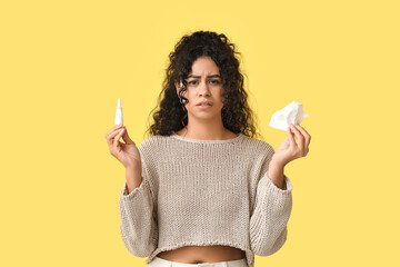 Young African-American woman with handkerchief and nasal spray suffering from allergy on yellow background