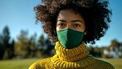 Young woman wearing a green mask and yellow sweater in a sunny outdoor park setting with trees in the background