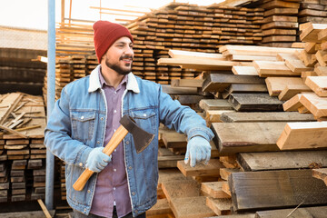 Fototapeta premium Handsome carpenter with axe near wooden planks at sawmill