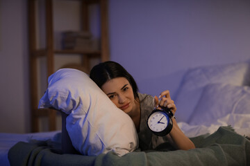 Beautiful young tired woman with alarm clock suffering from insomnia on bed in evening at home