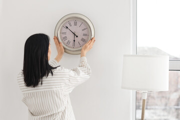 Young woman hanging clock on light wall at home, back view