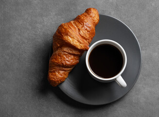 Cup of espresso coffee and fresh croissant on grey plate on dark background.