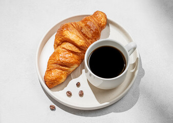 Cup of freshly brewed espresso coffee and fresh croissant on white plate on light background with shadow.