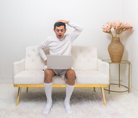 A man sits on a modern white couch, holding his head in surprise while interacting with his laptop in a minimalist living room.