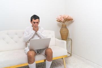 A man sits on a modern white couch, holding his head in surprise while interacting with his laptop in a minimalist living room.