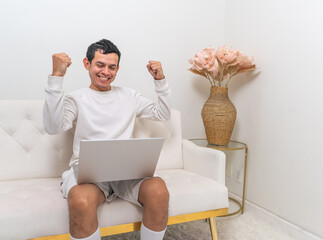 A man sits on a modern white couch, holding his head in surprise while interacting with his laptop in a minimalist living room. happy