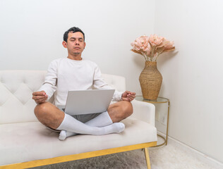 A man sits cross-legged on a modern white couch, practicing meditation with closed eyes while his laptop rests on his lap in a serene and stylish home setting.