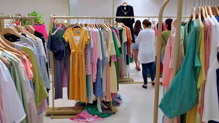 �Colorful mound of clothing in diverse sizes and styles dominates a spacious retail area, as shoppers browse, highlighting textile waste and fast fashion's impact.