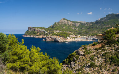 Fototapeta premium Lighthouse on rocky shore of Port de Soller, surrounded by turquoise waters of Srediterranean Sea, against backdrop of mountains and clear skies. Mallorca.
