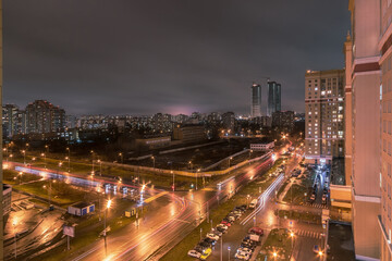 Fototapeta premium City lights twinkle against a cloudy night sky as traffic flows along roads below. Tall buildings dominate the skyline, highlighting urban life after dark.