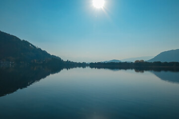 Breathtaking view of Lake Ossiach with water reflections of forest covered hills of Ossiacher Tauern in Carinthia, Austria, Europe. Clear blue sky in summer. Serene and idyllic scene in Austrian Alps