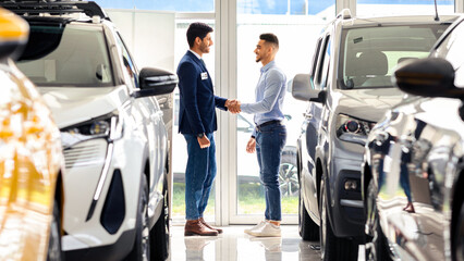Wealthy middle-eastern man standing among luxury cars in showroom and shaking sales manager hand, celebrating successful deal. Arab guy buying brand new comfortable auto in salon, full length