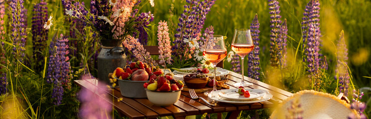 Banner. Countryside beautiful wedding decor, surprise romantic date outdoors, garden arrangement with lupins flowers on a meadow. Fruits, rose wine, burning candles. Sunset, candlelight, golden hour © ArtSys
