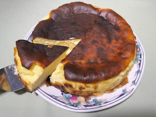 Closeup of a steel cake serving tool lifting up and pulling out the first triangular slice of a whole round, soft, baked cheesecake dessert on a plate.